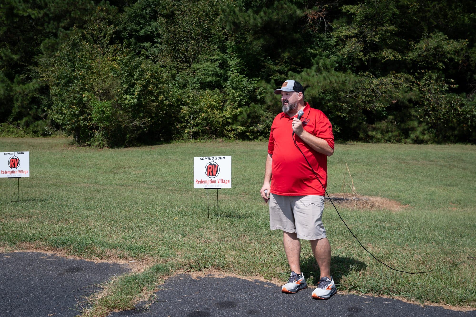 AIAO Ministry leader speaking at the Redemption Village groundbreaking, with “Coming Soon” yard signs on the future tiny homes site.