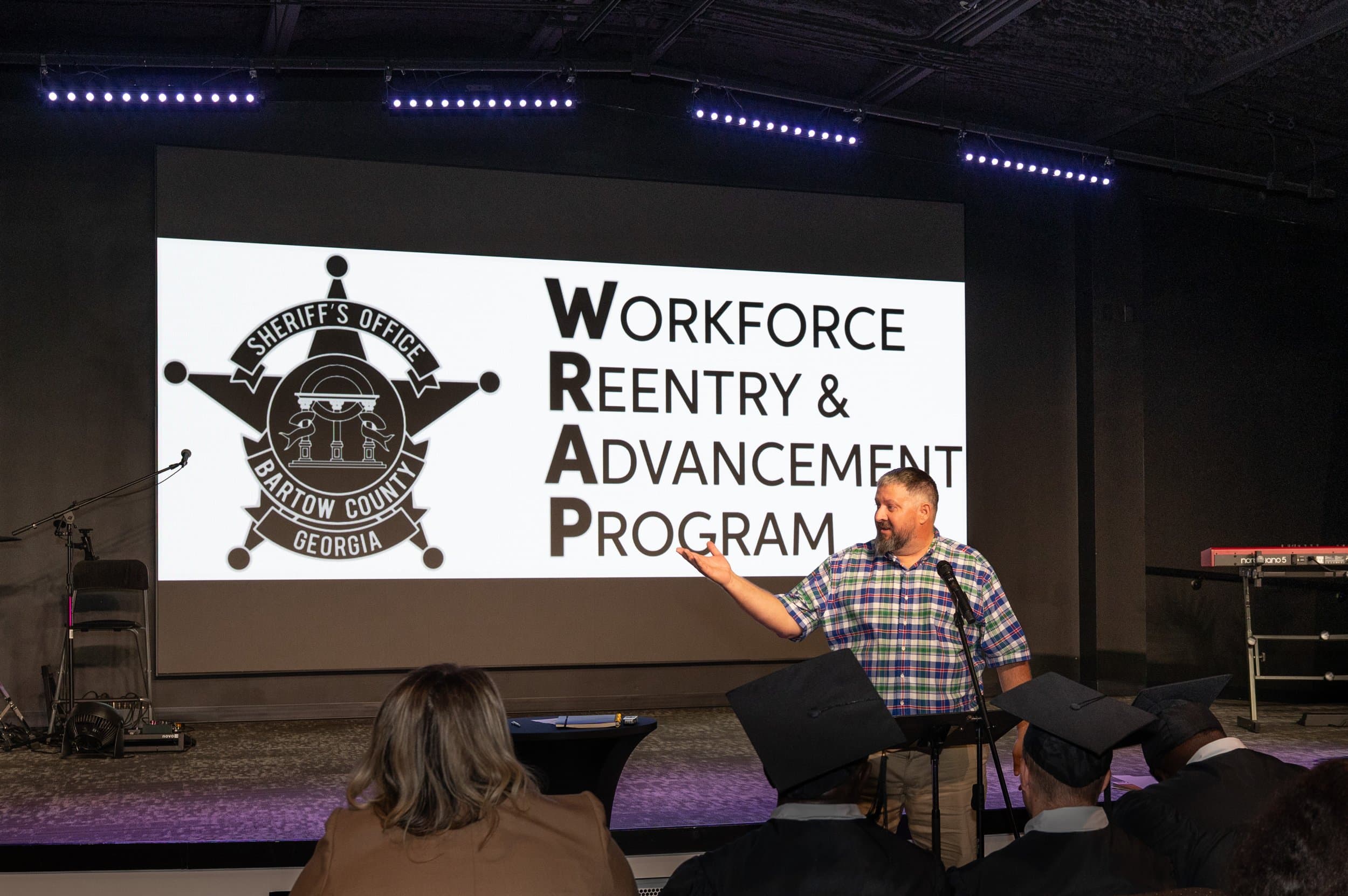 A man presenting the Workforce Reentry & Advancement Program to graduates and an audience